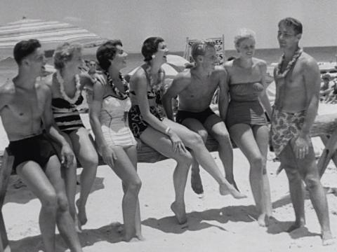 6 young men and woman in swimmers sitting on a tree trunk at the beach while another man speaks to them.