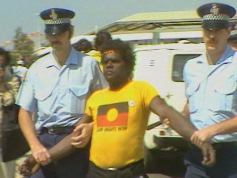 An Aboriginal protester wearing a yellow T shirt baring a Aboriginal flag is led away by two police officers