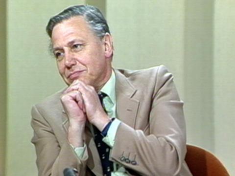 Naturalist and broadcaster David Attenborough clasps his hands underneath his chin in a listening pose. He is sitting in a studio.