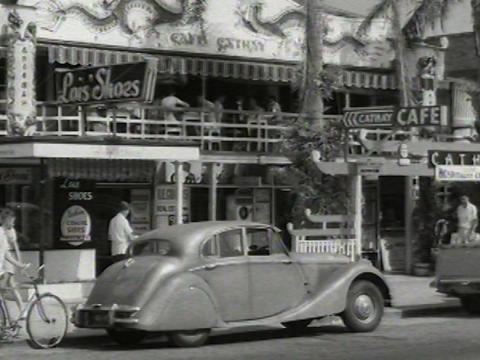 Street scene of a 1950s Daimler parked outside of shops.