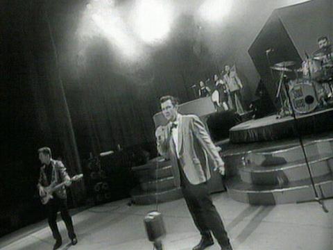 Jimmy Barnes, dressed in a tuxedo, is on stage singing into a microphone with a band behind him.