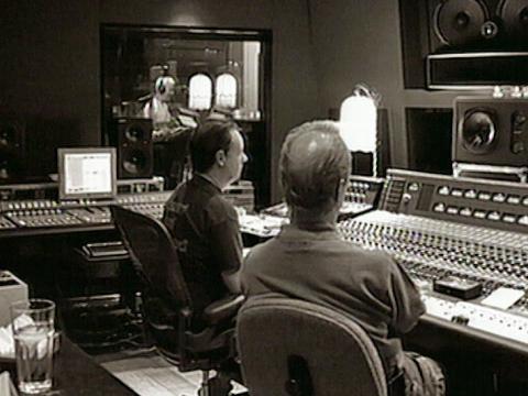Two men sitting behind the recording decks in a studio, in the far background through a window is Jimmy Barnes wearing headphones and singing into a microphone. 