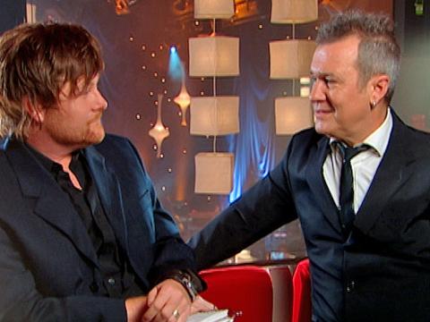 Music television host Chit Chat talks to singer Jimmy Barnes. The two men are both wearing black suits and facing each other. 