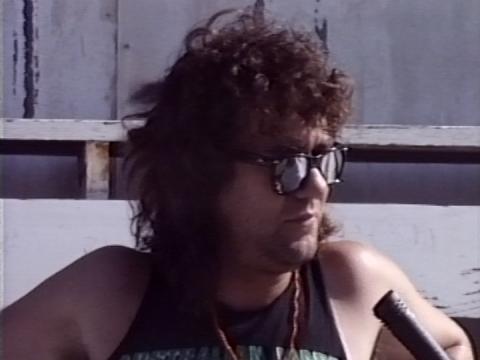Close up head and shoulders shot of Jimmy Barnes wearing sunglasses and a black singlet with a microphone directed at him. 