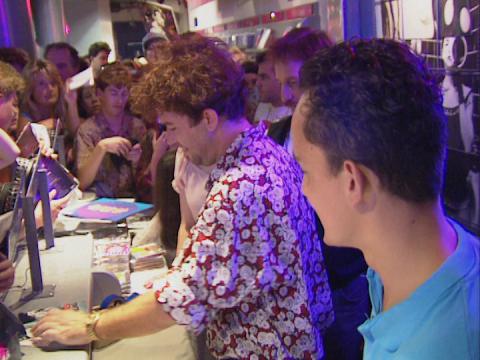 Singer Jimmy Barnes is standing behind a counter in a store signing autographs. He is surrounded by fans on the other side of the counter.