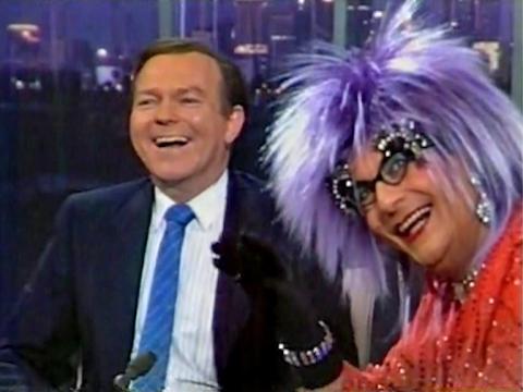 Mike Walsh laughs, looking off camera. In the foreground Dame Edna Everage waves off camera. She is wearing a wisteria coloured Tina Turner wig and glasses.