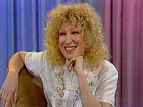 Comedian, singer and actress Bette Midler sits in a chair smiling with her chin resting on her hand.