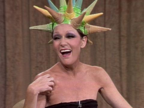 Jeanne Little laughs heartily. She is sitting in a studio and is wearing a hat made out of coloured ice cream cones.