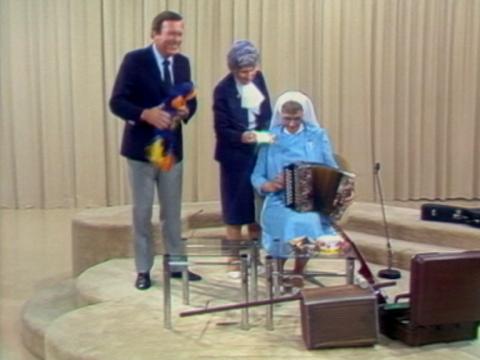 Mike Walsh, wearing a suit and tie, holds a colourful bird puppet. A nun in a blue habit is seated and she holds a piano accordian. A woman stands behind her holding a harmonica.