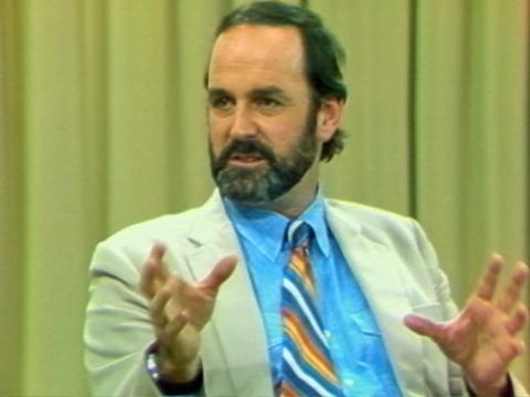 Comedian John Cleese talking and gesticulating with his hands. He is wearing a suit and tie.