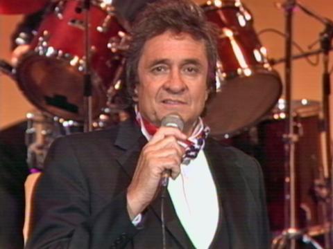 American singer Johnny Cash sings with a microphone as he looks directly at the camera.