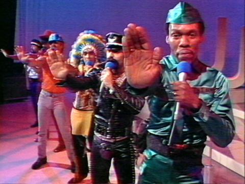 The six members of the disco group The Village People stand in a line. They al hold their hands out in a stop motion with microphones in their other hand. They all wear colourful character costumes.