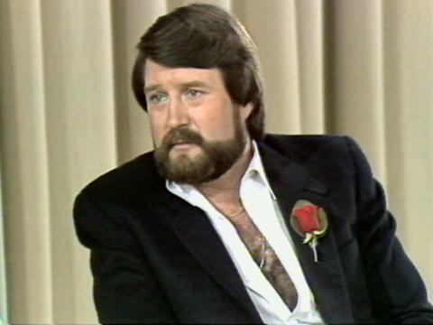 Journalist and politician Derryn Hinch is sitting in front of a curtain. He has a red rose on his lapel.