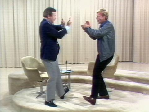 Mike Walsh and actor Jack Thompson are doing tai chi. They have their legs bent slightly, feet out and arms in the air in front of them.