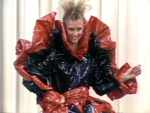 A woman with very 80s spikey hair is wearing a plastic shiney dress with massive frills.
