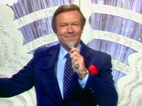 Mike Walsh stands with a microphone in his hand in front of a sparkly curtain.