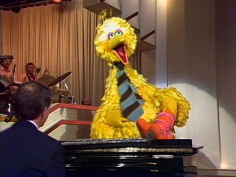 The puppet Big Bird is sitting on a piano with his feet up. He's wearing a tie.