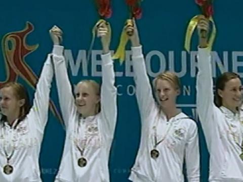 Four Australian women's swimmers stand on the gold medal podium, arms raised holding bouquets.