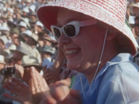 A women in a large pink had an white retro sunglasses claps excitedly in a crowded stadium
