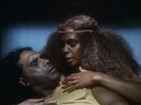 A man in a jail cell is cradled by a woman in traditional Aboriginal Australian ceremonial costume