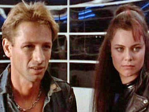 A man in a denim shirt and a woman in a leather jacket stand in front of a futuristic background