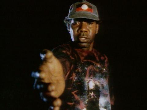 A man wearing an Australian Aboriginal Flag hat reaching a hand towards the viewer in front of a black background