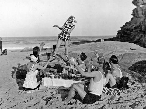 A group of men and women sitting around a small table on the sand at a beach while a woman on a large rock dances in front of them.