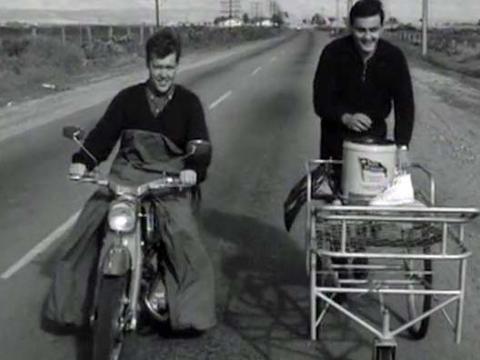 A man pushing a hospital bed down a road with a motorbike rider next to him.