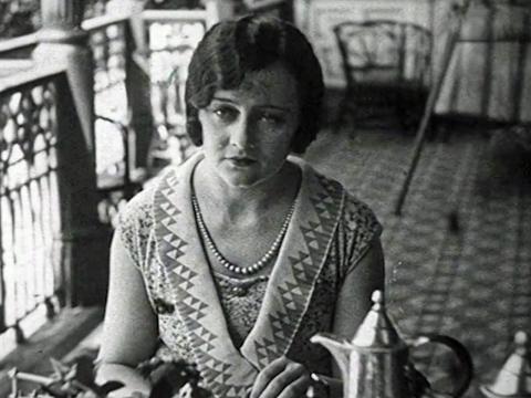 Actress Isabel McDonagh (billed as Marie Lorraine) in a scene from The Far Paradise. She is pictured from the waist up wearing a 1920s style dress. She is seated at a table and looking forlorn.