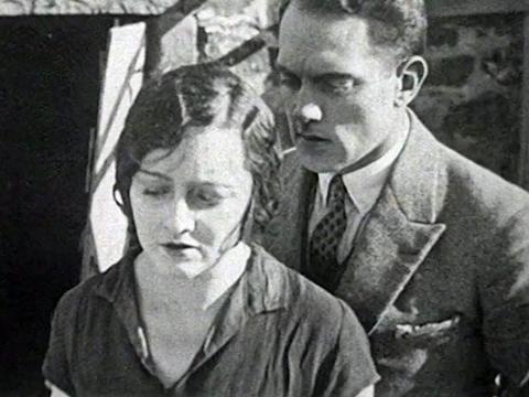 Actress Isabel McDonagh (billed as Marie Lorraine) in a scene from The Far Paradise. She is pictured from the shoulders up facing towards the camera and looking sad as a man (actor Paul Longuet) in a suit stands closely behind her speaking into her ear.