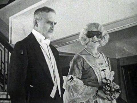 An elderly man and young woman pictured from the waist up, dressed for a masked costume ball, circa 1928.