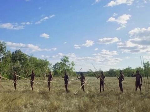 A line of Aboriginal men wielding spears on a grass plain under a sunny blue sky