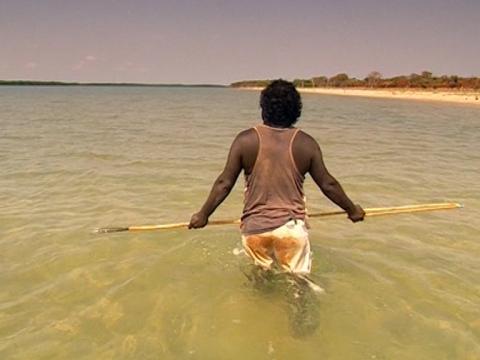 An Indigenous man walking through the water holding a spear