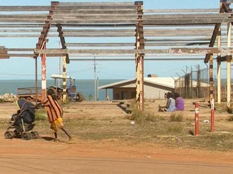 Indigenous woman pushes a pram at a settlement in Arnhem Land, other Indigenous people and the sea in the background