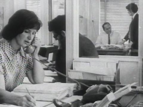 In the foreground is a woman sitting at a desk writing and speaking on the phone, other office workers in background