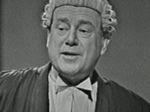 A prosecutor (Kenric Hudson) wearing a barrister's wig