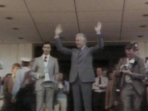 Prime Minister Gough Whitlam standing on the steps of Parliament House with his hands raised, surrounded by party members, police and press, Paul Keating stands next to him holding a megaphone