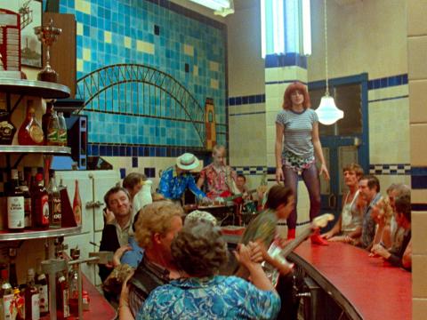 A woman stands on the bar of the Harbour View Hotel as she sings a song with a live band. The wall behind them has a tile mosaic of the Sydney Harbour Bridge.