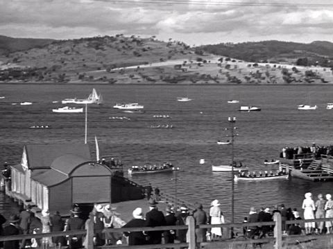 A black and white image of boats and a rowing regatta on the Derwent River. Onlookers watch from the riverbank.