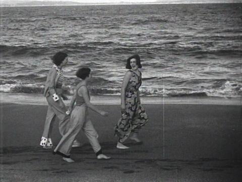 Three women walking along a beach, the woman in front turns laughing at her companions behind her