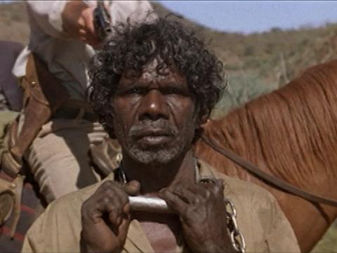 The tracker (David Gulpilil) stands holding the chain around his neck as a man behind on horseback points a gun at him