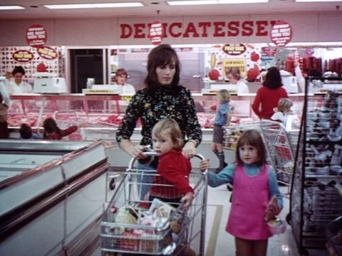 A woman pushes a supermarket shopping trolley underneath a big sign that says 'delicatessen'. She has a small child sitting in her shopping trolley and an older child walks along next to the trolley.