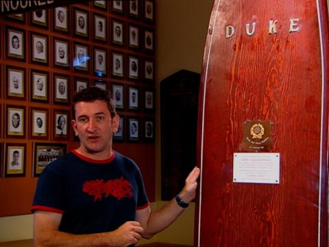 Warren Brown stands next to a long wooden surfboard with the word 'Duke' written on it. 