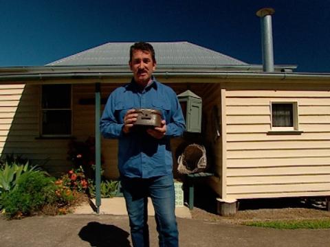 Warren Brown stand outside a small weatherboard building holding a small box.