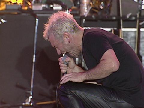 Jimmy Barnes kneeling down on stage singing into a microphone. 