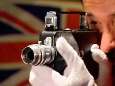 Close up of Warren Brown wearing gloves and looking through the lens of an older style movie camera.