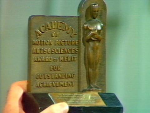 A hand holds a statue inscribed 'Academy of Motion Picture Arts and Sciences Award of Merit for Outstanding Achievement'.