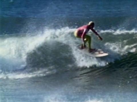 Australian surfing champion Pam Burridge in the surf, riding a wave on a surfboard, wearing a pink and yellow wetsuit