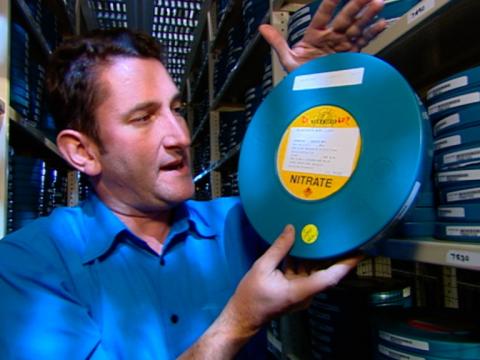 Warren Brown in an archive holding a round tin that contains a print of the film 'The Sentimental Bloke'.
