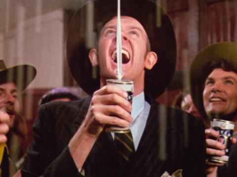 Barry Crocker as Barry McKenzie opens a can of Fosters beer and the beer shoots up in the air above and rains down on him and the others around him. They are all wearing digger's hats.